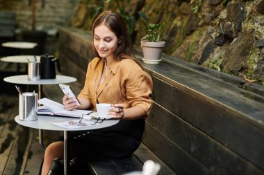 Kafedeki genç bir kadın, akıllı telefonunu kullanarak kahvesinin tadını çıkarıyor ve günlük hayatını özgüvenle kucaklıyor..