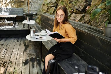 Sitting in a cozy cafe, a young woman with a prosthetic leg immerses herself in a notebook, near cup of coffee. clipart