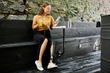Protez bacaklı genç bir kadın bir kafede rahatlar, bir içki yudumlar ve telefonunu kontrol eder, günlük hayatı somutlaştırır..