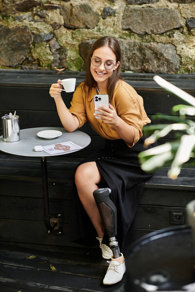 В очаровательном кафе молодая женщина с протезной ногой улыбается, потягивая кофе и проверяя телефон, сливаясь с повседневной жизнью.