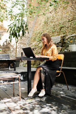Genç bir kadın bir kafe masasında oturuyor, dizüstü bilgisayarına odaklanmış, protez bir bacak ve sıcak bir atmosferle hayatı kucaklıyor..