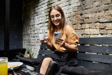 Yapay bacaklı genç bir kadın kahvesini yudumlarken gülümsüyor ve büyüleyici bir kafede telefonunu karıştırıyor.