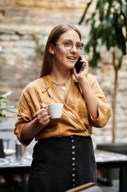 Genç bir kadın bir fincan kahve içerken telefonda sohbet ediyor, canlı bir kafe atmosferini somutlaştırıyor..