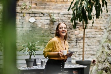 Genç bir kadın bir fincan kahve ve bir telefon tutuyor, sıcacık bir kafe bahçesinde sıcacık gülümsüyor.