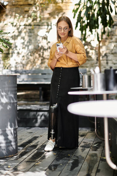 Молодая женщина с протезной ногой стоит в кафе, взаимодействуя с телефоном в окружении растений.