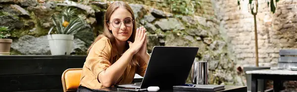 stock image A young woman sits at a cafe, focused on her laptop in a garden setting.