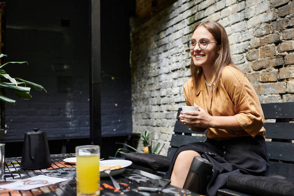 Молодая женщина удобно сидит в кафе, радостно улыбаясь, держа чашку, подчеркивая свой яркий образ жизни.