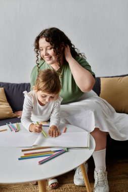 Bir anne, kızının küçük bir masada parlak pastel boyalarla resim yapmasını zevkle izler..