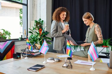 Two coworkers engage enthusiastically while discussing work amid vibrant LGBT decorations. clipart