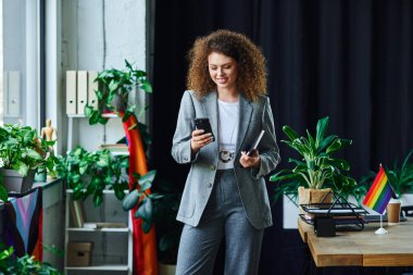 A smiling colleague connects with friends on her phone in a vibrant, plant filled workspace. clipart