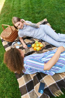 İki arkadaş bir battaniyenin üzerinde rahatlar, hikayelerini ve lezzetli meyvelerini açık mavi gökyüzünün altında paylaşırlar..