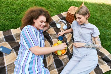 Two friends share laughter and snacks on a blanket, soaking in the sun during their picnic. clipart