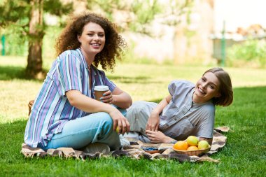 Two friends relax on a blanket outdoors, sharing smiles and enjoying delicious snacks together. clipart