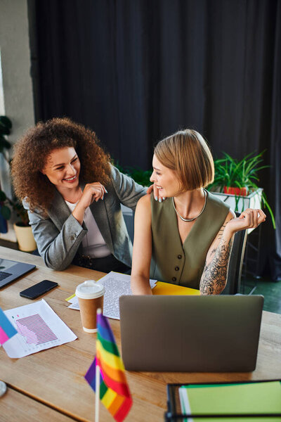 Two coworkers chat animatedly amid vibrant flags of the LGBTQ community.