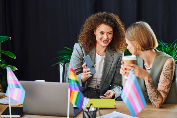 Two coworkers engage joyfully at their desks, surrounded by pride flags and positive energy.