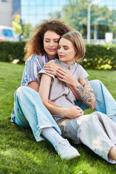 Two young women sit close in a grassy park, sharing moments of joy and connection.