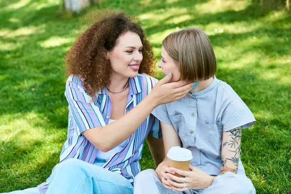 stock image Two young women enjoy a cheerful moment under the sun while sipping coffee in the park.