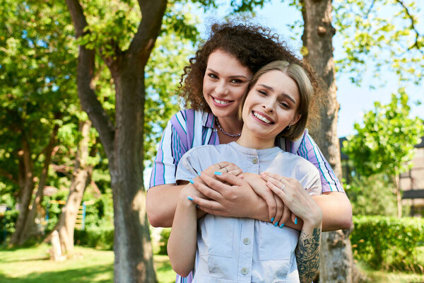 Two friends share joyful moments outside, embracing in a lively park filled with trees.