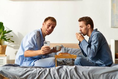 İki gülümseyen adam pijamalarıyla kahve ve hamur işlerinin tadını çıkararak birlikte keyifli bir sabah anı yaratıyorlar..