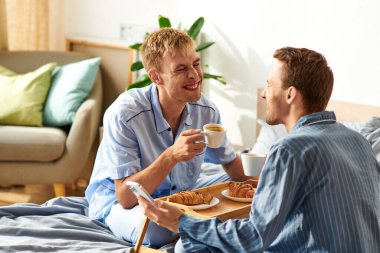 Two men in pajamas enjoying breakfast together, sharing laughter and affection in their bright bedroom. clipart