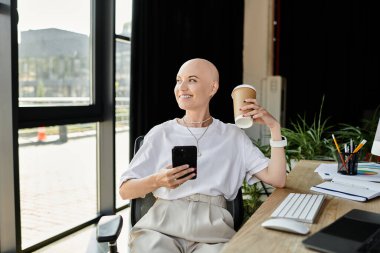 Genç kel bir kadın zarif bir şekilde gülümseyerek iş yerinde kahve ve akıllı telefonunu dengeliyor..