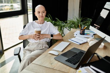 Kahvesini yudumlayan kel bir kadın yeşil ve teknolojiyle çevrili masasına yansıyor..