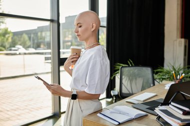 Zarif giyinmiş genç kel bir kadın, şık bir çalışma alanında telefonunu kontrol ederken kahvenin tadını çıkarıyor..