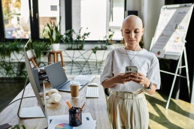 A young bald woman dressed elegantly stands in a chic office, focused on her phone. clipart