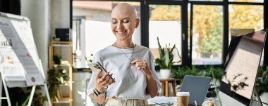 Zarif bir şekilde giyinmiş genç kel bir kadın şık bir ofis ortamında telefonuna gülümsüyor..