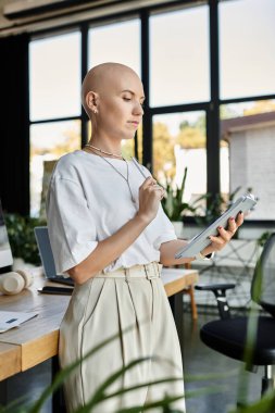 Şık giyinmiş genç kel bir kadın çalışma alanında derin düşüncelere dalmış bir tabletle meşgul oluyor..