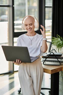 A young bald woman dressed elegantly enjoys music and interacts with her laptop in a bright office. clipart