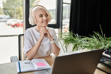 Genç, kel bir kadın, parlak ve modern bir ofiste dizüstü bilgisayarıyla çalışırken gülümsüyor..