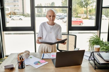 Şık giyinmiş genç kel bir kadın çalışma odasındaki dosyalarını inceliyor..