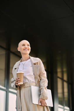 Şık giyinmiş genç kel bir kadın, elinde bir kahve fincanı ve dizüstü bilgisayarla güvenle duruyor, gülümsüyor..