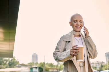 Nazikçe giyinmiş neşeli genç bir kadın telefonuyla sohbet ederken elinde kahve fincanı tutuyor..