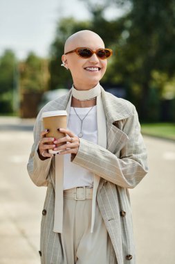 A young bald woman radiates confidence in elegant attire while holding coffee outdoors. clipart