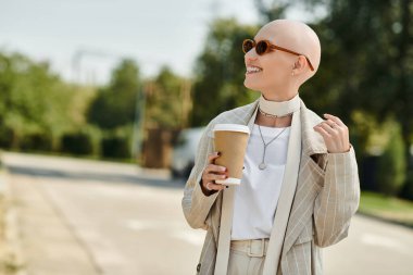 With a joyful expression, a bald woman in stylish clothing holds her drink in bright daylight. clipart