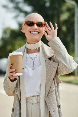 Güneş gözlüğü takan ve zarif kıyafetler giyen neşeli, kel bir kadın dışarıda kahvesinin tadını çıkarıyor..