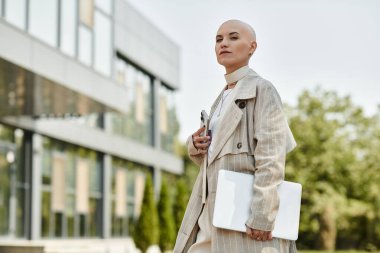 Zarifçe giyinmiş genç kel bir kadın dizüstü bilgisayar ve kalem tutuyor..