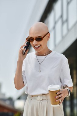 Şık giyinmiş, kel bir kadın telefonda konuşurken gülümseyerek kahveyi tutuyor..