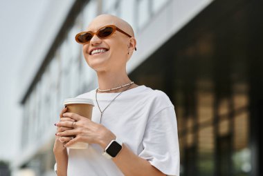 Kel kafalı genç bir kadın dışarıda şık kıyafetler içinde kahvenin tadını çıkarırken ışıl ışıl gülümsüyor..