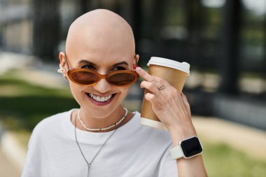 Genç ve kel bir kadın elinde kahve fincanı tutarken zarif bir şekilde gülümsüyor ve gününün tadını çıkarıyor..