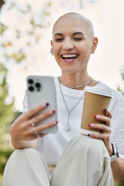 Neşeli, kel bir kadın zarif bir şekilde kahvesini yudumluyor ve gün ışığında telefonuna gülümsüyor..