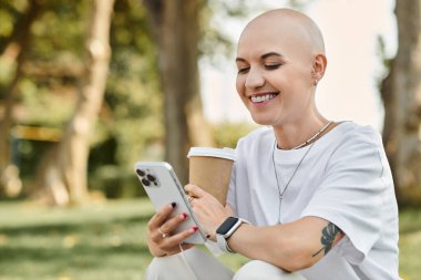 A cheerful bald woman dressed elegantly sips coffee while engaging with her phone outdoors. clipart