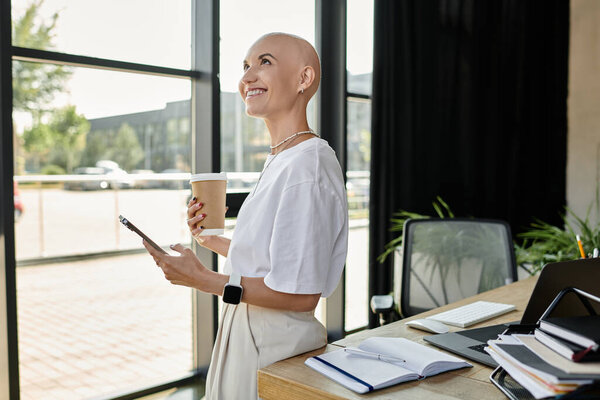 Молодая лысая женщина в изысканной одежде смотрит в окно с теплой улыбкой, держа в руках кофе.