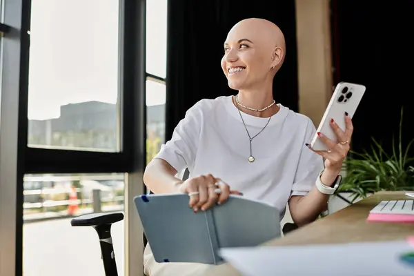 stock image A cheerful young woman in elegant attire sits at her desk, engaged with her smartphone and notebook.