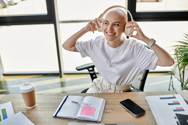 Молодая лысая женщина в стильной одежде слушает музыку с улыбкой, сидя за столом.
