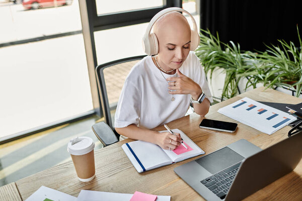Молодая лысая женщина вдумчиво пишет заметки в наушниках на своем стильном рабочем месте.