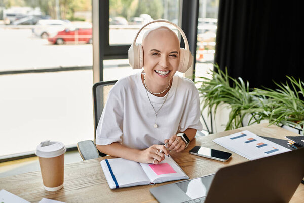 Молодая женщина, лысая и элегантно одетая, радостно занимается своей работой за столом.