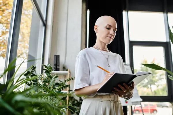 stock image A young bald woman in elegant attire is thoughtfully writing in a notebook, surrounded by greenery.